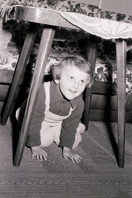 A young child crawls under a dark wood table with a patterned tablecloth, appearing to play hide-and-seek. The child wears a ...