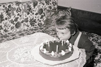 A young child, likely a boy, blows out candles on a chocolate cake, seated before a lace-covered table. Floral patterned upho...