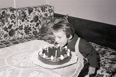 A young child, likely a boy, blows out candles on a chocolate cake adorned with cherries, seated on a floral-patterned sofa. ...