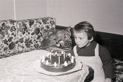 A young child, possibly a boy, stands observing a birthday cake with lit candles. The scene suggests a domestic setting, like...
