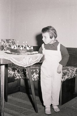 A young boy stands beside a table laden with a birthday cake, adorned with lit candles. He wears checked overalls over a dark...