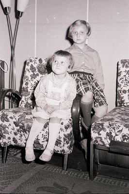Two young girls seated in patterned armchairs, captured in a candid black and white snapshot. One wears a dress and knee sock...