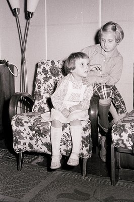 A young girl in a floral-patterned dress and tights sits in an upholstered armchair, with another child standing behind her. ...