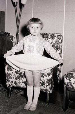 Young girl playfully holds up a dress, showcasing a frilled hem. She wears a patterned top, suspendered skirt, knee socks, an...