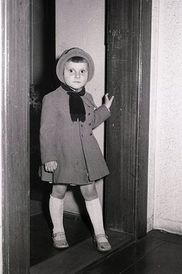 A young child, likely a girl, stands in a doorway wearing a wool coat, cloche hat, and saddle shoes with stockings. The scene...