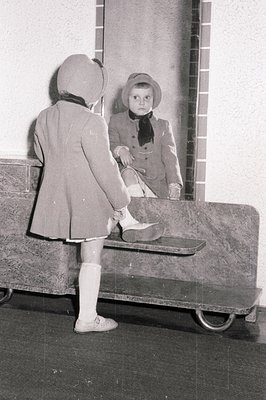 Young child in a double-breasted coat, hat, and knee socks, captured in a mirror reflection. Appears to be dressing or adjust...
