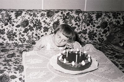 A young boy with a bowl cut smiles next to a chocolate cake with eight lit candles. He reclines against a floral-patterned so...