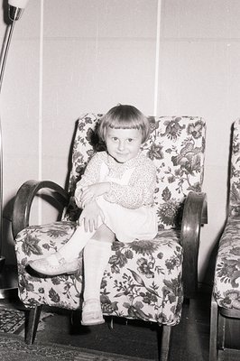 A young child sits formally in a patterned armchair, appearing to pose for a portrait. The room features paneled walls and a ...