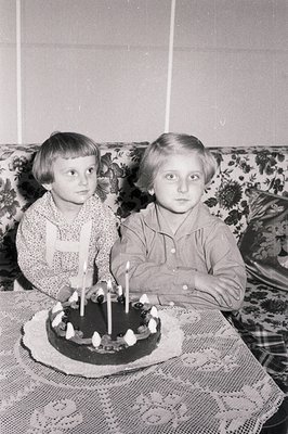 Two young children, a boy and a girl, sit side-by-side with a birthday cake featuring lit candles. The girl wears a collared ...