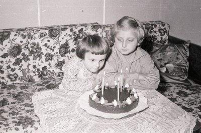 Two young girls celebrate a birthday with a chocolate cake adorned with seven candles. The scene appears domestic, set agains...