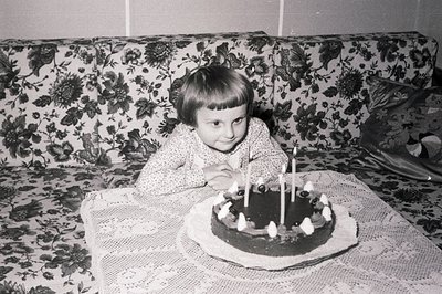 A young child, likely a girl, sits poised before a birthday cake with eight lit candles on a lace-covered table. The backdrop...