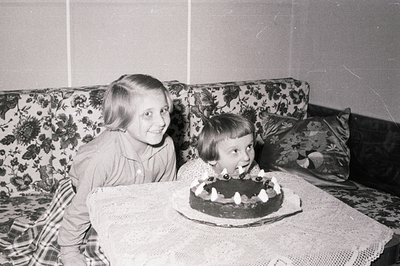 Two young girls share a moment of joy, excitedly observing a chocolate cake with lit candles. The setting appears to be a hom...