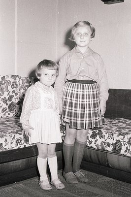 Two young girls stand near a floral-patterned couch. One wears a white, short-sleeved dress with lace trim; the other, a plai...