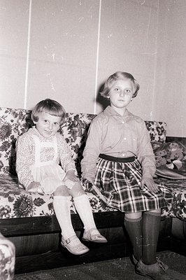 Two young girls, seated on a floral upholstered bench, pose for a formal portrait. Both wear dresses and knee-high socks; one...