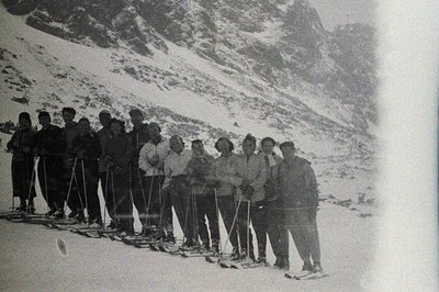Group portrait of thirteen individuals, likely skiers, posed on a snow-covered slope with a dramatic mountain backdrop. Cloth...