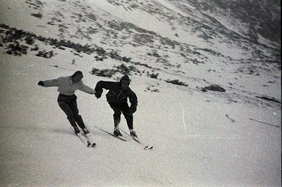 Two skiers descend a snow-covered slope, likely in an alpine region. The image, captured with a vintage film camera, exhibits...