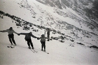 Three skiers descend a snow-covered slope. Visible clothing suggests a mid-century style. The backdrop features a snow-dusted...