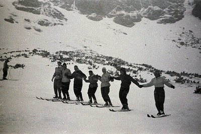 A group of seven people, all wearing winter clothing and standing on skis, hold hands in a circle on a snow-covered slope. Mo...