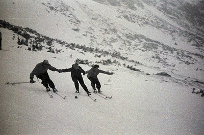 Three skiers descend a snow-covered slope. The individuals wear period clothing, with dark jackets and trousers. Visible terr...