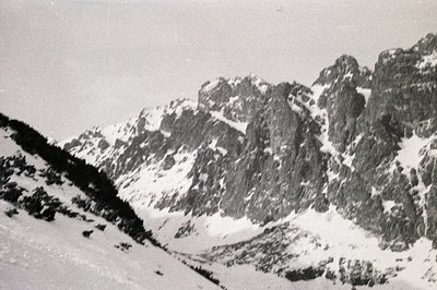 Dramatic, snow-covered alpine landscape featuring steep, rocky peaks. Sparse vegetation clings to the lower slopes. Likely a ...