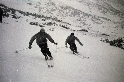 Two skiers descend a snow-covered slope, captured in a vintage black and white photograph. The men wear heavy winter clothing...