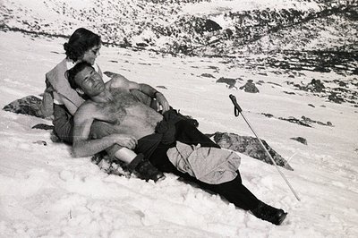 Man lies prone in snow, partially unclothed, supported by a woman. Likely an alpine environment, judging by the snow-covered ...