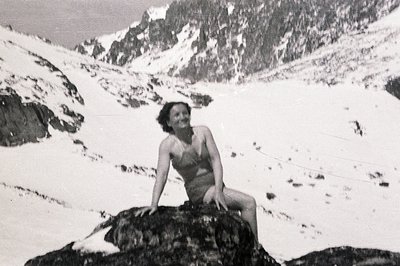 A young woman in a vintage swimsuit sits perched on a rock amidst a snowy, mountainous landscape. Likely a promotional shot s...