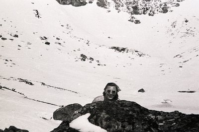 A man with long hair smiles directly at the camera while partially sheltered by dark rocks covered in snow. Sparse vegetation...