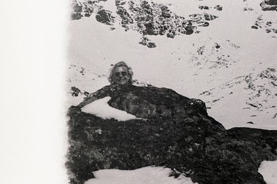 A man with a full beard sits atop a snow-covered boulder, gazing toward a mountainous, snowy landscape. The image, likely a p...