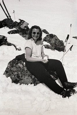 A woman sits on a boulder amidst a snowy landscape, wearing a short-sleeved white shirt, dark trousers, and sunglasses. Likel...