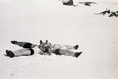 Three figures lie intertwined in a snowdrift, appearing relaxed or playfully entangled. Their attire - shorts & boots - sugge...