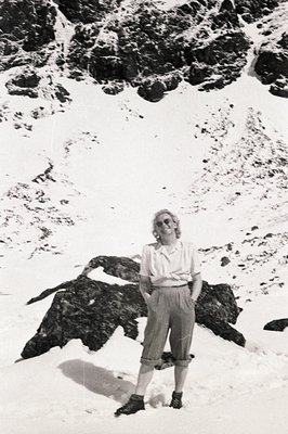 A woman in 1960s-era clothing stands confidently amidst a snowy, rocky alpine landscape. She wears wide-leg trousers, a butto...