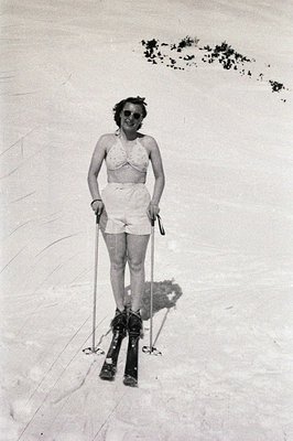 A woman in a vintage swimsuit and ski boots poses on snow. She wears sunglasses and holds ski poles, appearing poised. Likely...
