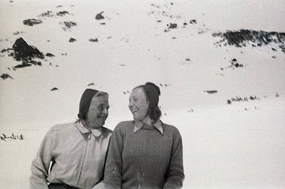 Two women with smiling expressions, bundled in warm winter clothing & hats, pose outdoors in a snowy landscape. Likely a cand...