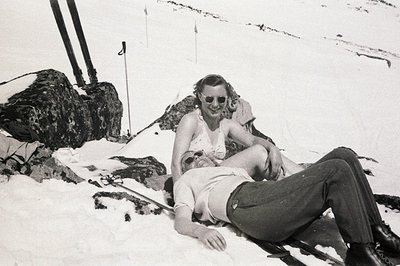 A man and woman rest on a snowy slope, reclined against a large rock formation. The woman, wearing a sleeveless dress and sun...