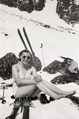 A poised woman in a one-piece swimsuit sits on a snowbank, legs crossed, with vintage skis and poles nearby. She wears round ...