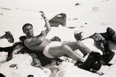 A man, shirtless and in shorts, reclines on a snow-covered rock formation. He appears to be posing with an air of relaxed con...