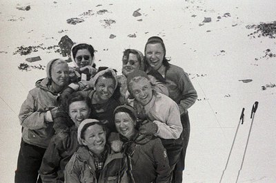 A group of women, bundled in warm outerwear, pose outdoors in a snowy landscape. Smiling faces, knit hats, and vintage ski eq...