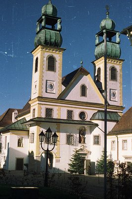 Grand Baroque-style church facade features twin towers topped with green domes. The structure includes intricate detailing, s...