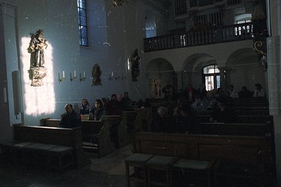 A view inside a church. Several people are seated in pews, observing an event or service. Stone statues and architectural det...