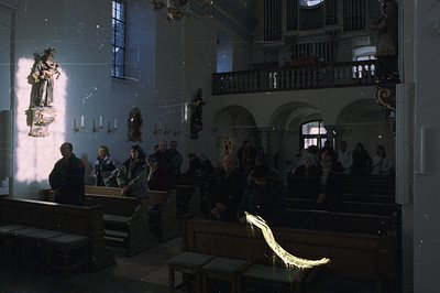 A dimly lit, historic church interior. Several figures are seated in pews while others stand or are visible on a balcony. A s...