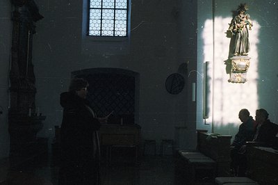 Interior of a stone church, featuring a dark wooden pulpit, simple benches, and a statue of a robed figure illuminated by sun...