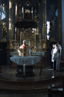 A liturgical scene captures a priest celebrating mass before an ornate, gilded altar. Side chapel statues and stained glass i...