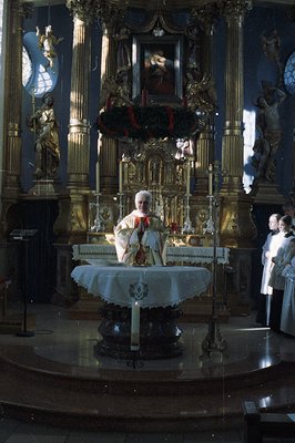 A Catholic priest, likely celebrating mass, stands behind an ornate altar with gilded carvings. Statues of angels flank the s...