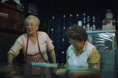Two women in aprons, likely shopkeepers, work behind a display counter filled with candies. Festive decorations, including a ...