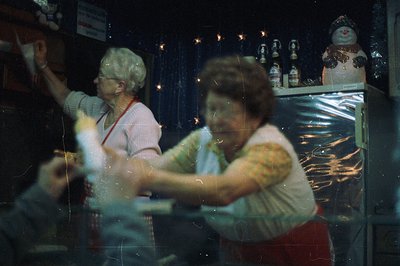 Two women in aprons work behind a counter, likely in a confectionery or gift shop. Visible merchandise includes figurines, de...