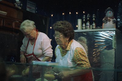 Two women in work aprons prepare food behind a stainless steel counter. A display of ornaments and decorations hangs on the w...