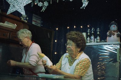 Two older women in aprons work behind a counter, likely preparing food or beverages. Christmas decorations – a snowman, twink...