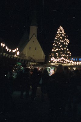 A community gathers at a nighttime Christmas market. A simple, peaked-roof building flanks a brightly lit, artificial fir tre...