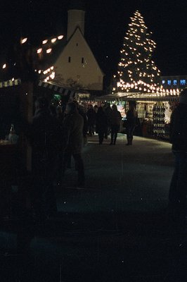 A brightly lit, snow-covered outdoor ice rink with a large, decorated Christmas tree. Figures in winter clothing skate around...
